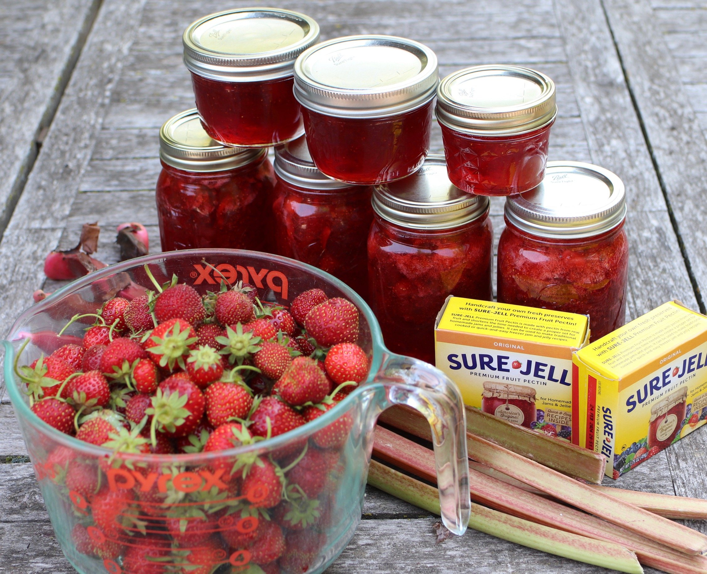 a bowl of strawberries, rhubarb stalks, pectin, and jars of jam